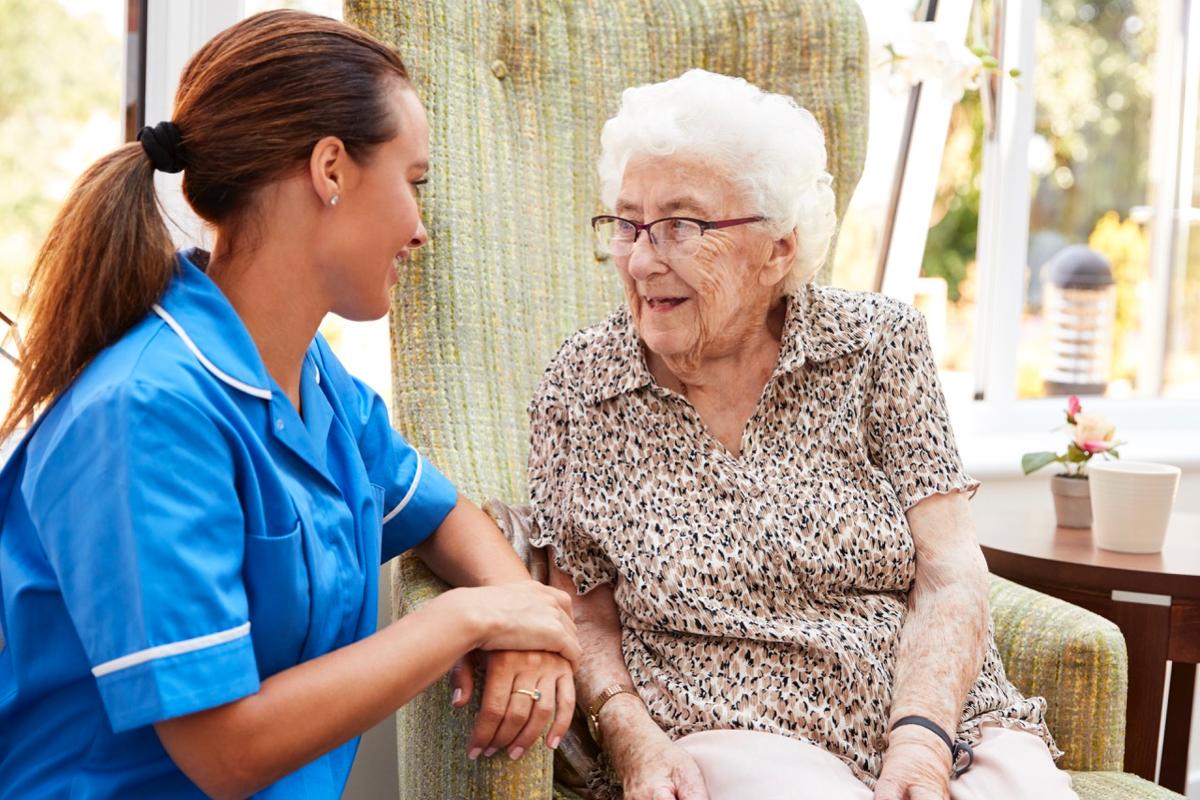 A health care worker in bright blue scrubs and a ponytail sits next to an old bespectacled Caucasian woman in a patterned collared shirt.