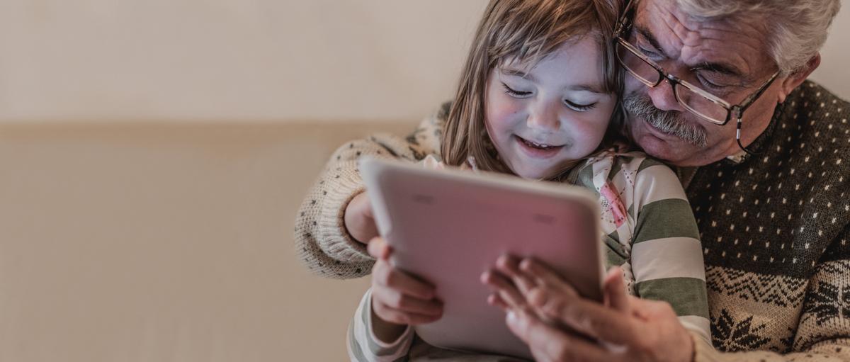 Grey-haired man in patterned sweater helps young Caucasian child with a tablet