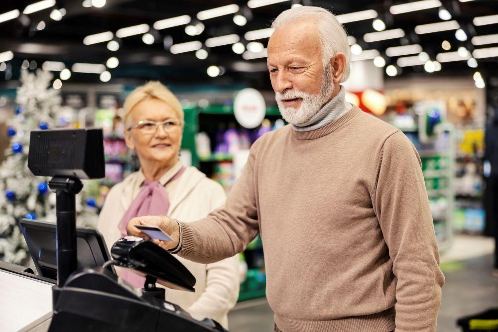 Image of a senior male and female at a store cash register. They both have an amused expression on their face, smiling. The male is swiping his debit or credit card on the cash register.