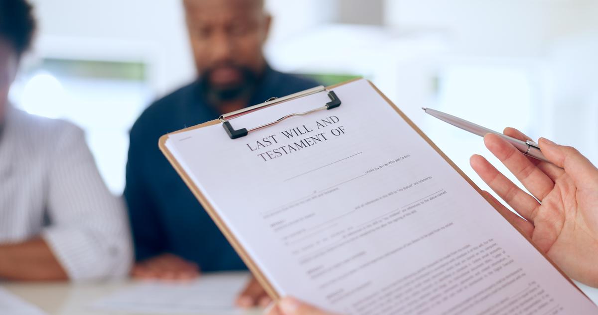 A document is sitting on a clipboard that reads Last Will and Testament Of. The person holding the clipboard is talking with a couple that is seated in the background. The man in the background is wearing a blue shirt and the person sitting next to him is wearing a white shirt. The couple look like they are reading documents that are placed on the table in front of him. 
