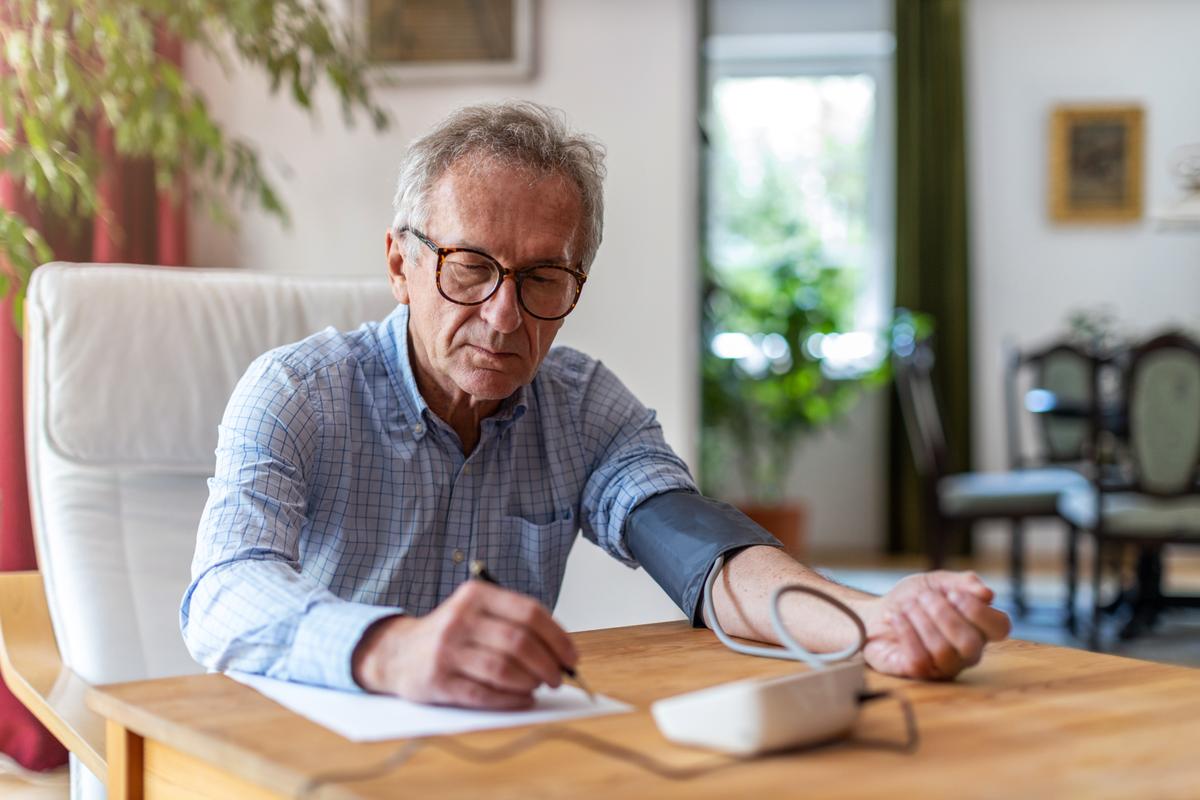 Man sitting at home taking his blood pressure. He is using an at home blood pressure machine with the cuff around his arm. He seems to be writing down what his results are. 