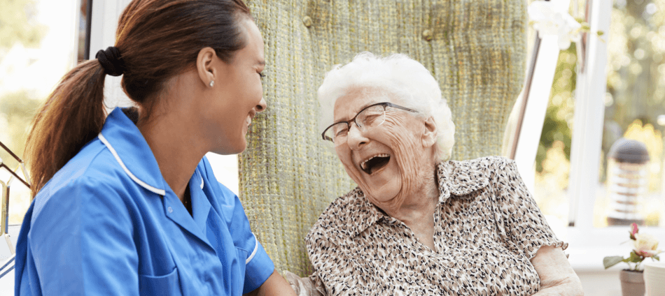 A senior woman with eye glasses laughing alongside her care giver who is wearing a blue uniform, they are in an outdoor area.