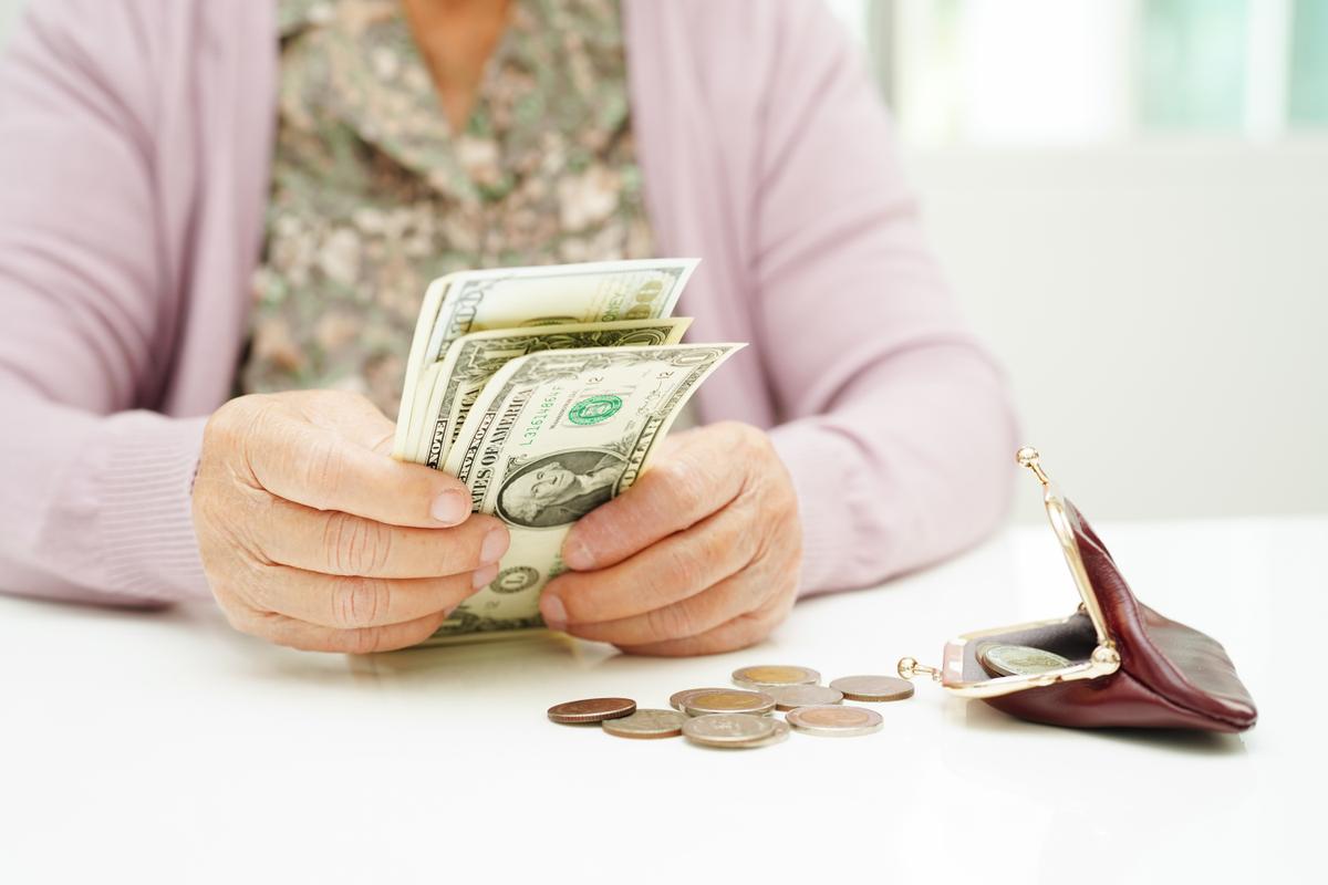 An elderly woman with a light pink sweater and floral top sitting at a table counting USD money. Her coin purse is open and there is some change on the table. 