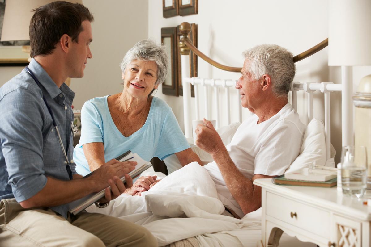 A doctor with a stethoscope and clipboard sits at the edge of an elderly couple’s bed in their home. The senior male is the patient laying in bed with a drink in his hand, while his wife is seated at the other edge of the bed.