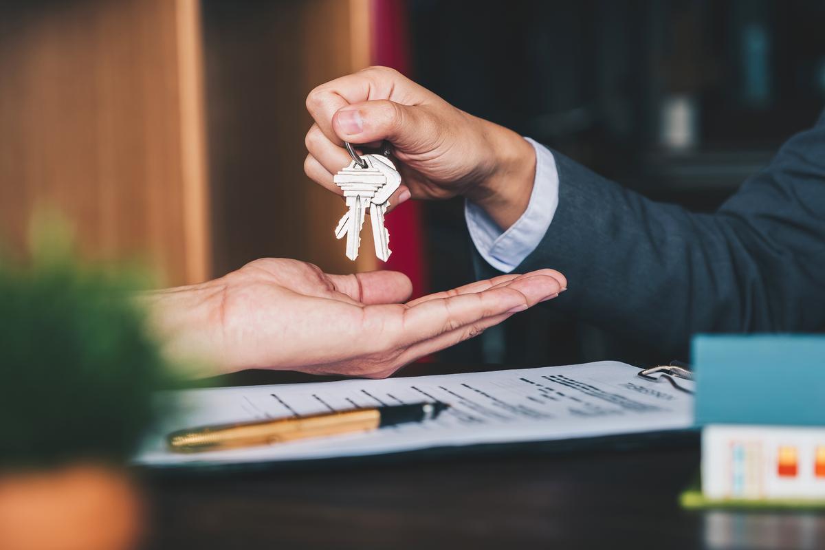 Two hands are seen. From the left side a hand is reaching out to grab a pair of keys that the right side hand is dangling over the left person's hand. There is paperwork on the table.