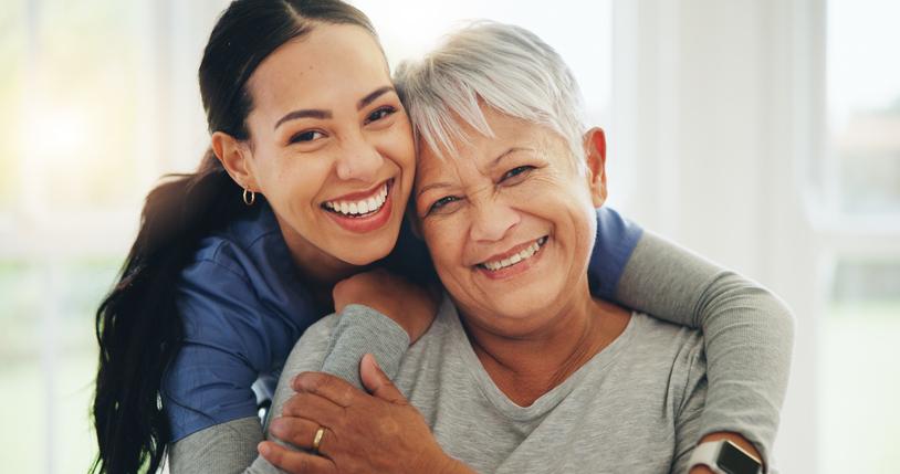 Smiling young female doctor, nurse, or caregiver with long hair in a ponytail and blue scrubs, hugging a smiling, elderly woman with short, white hair from behind while she is sitting in a chair.