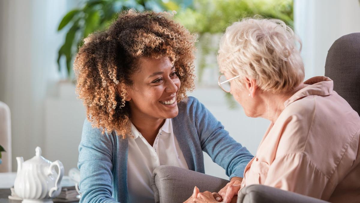 A senior woman receiving care while sitting on a gray couch, The caregiver is smiling and greeting her ready to offer a cup of tea. The tea mug is on a table behind both of them.