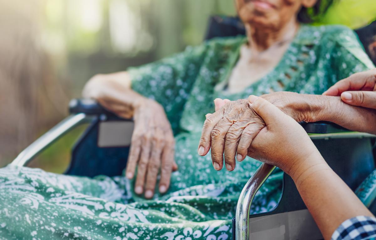 Closeup image of a senior woman sitting outside on a wheelchair wearing a green dress, there is a caretaker nearby holding her hand beside her.