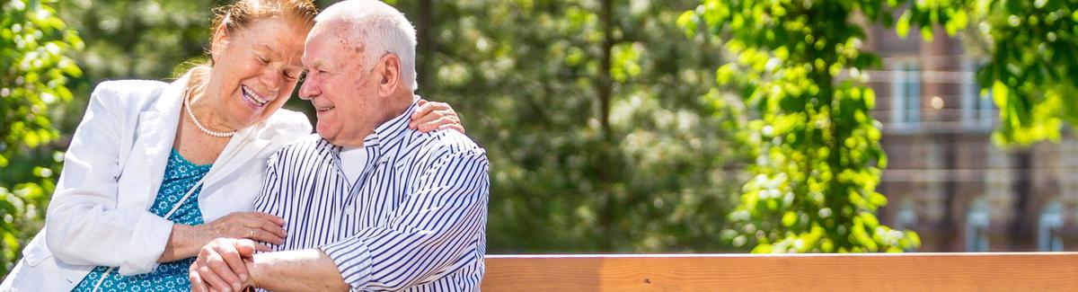 An elderly couple, man and woman, sit together on a bench in a local park. The couple are touching foreheads and smiling playfully with one another. The older gentleman is holding a cane.