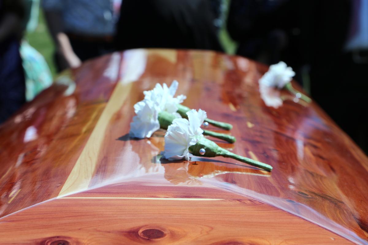 A closed brown casket with three small white flowers on top. The background shows a few people surrounding the casket as if this is at an outdoor funeral.