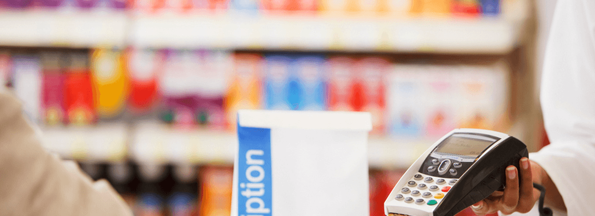 A person in a tan coat pays for their prescriptions at the pharmacy register. The pharmacist, a woman of color wearing a white lab coat, holds out a credit card reader to the person in the tan jacket.