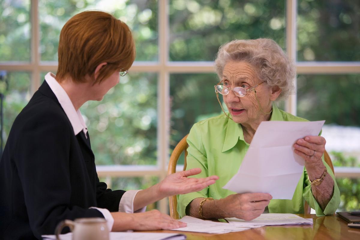 A senior woman is having a meeting with an attorney at her home. The attorney is telling the senior woman something while the senior woman is holding paperwork in her hand. 