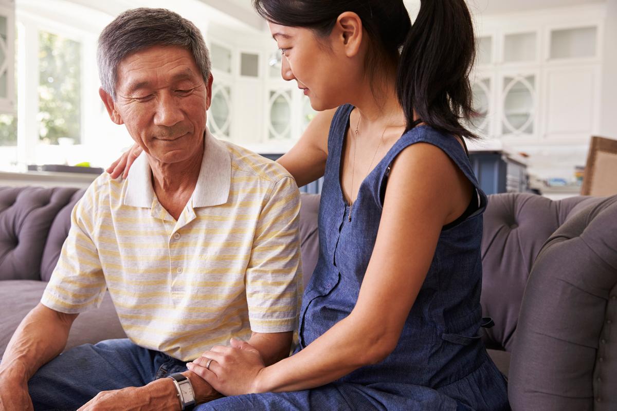 Image of a daughter and father sitting on a couch. The father is older and appears confused. The daughter has her arm around his shoulder comfortingly.