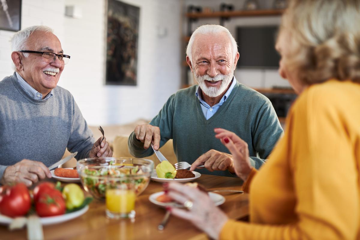Image of three seniors enjoying a healthy meal together around a dinner table, smiling, laughing, and socializing. 