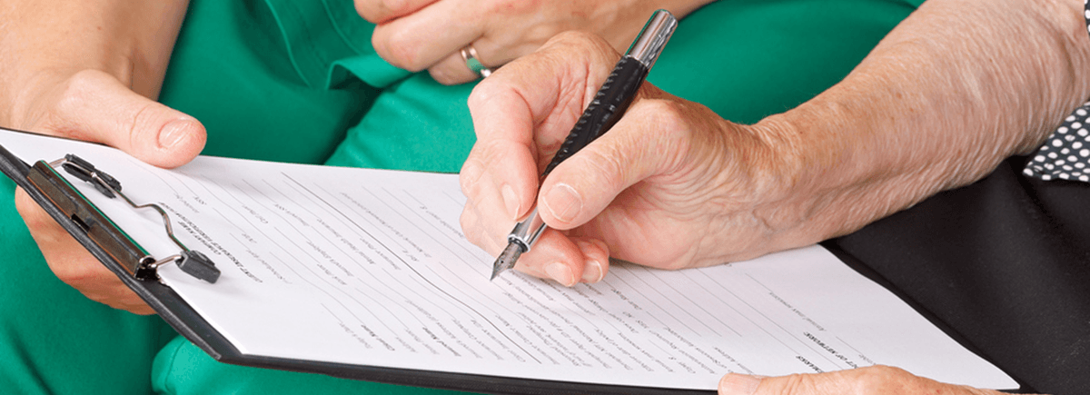A woman's hand is featured filling out forms with a pen. She is sitting next to another person who is wearing teal scrubs. You do not see their faces, you can only see their hands with the clipboard.