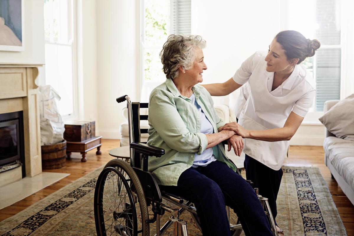 A woman with short, greying hair sits in a wheelchair in front of a fireplace. She wears a pale green linen button-down and smiles as she talks to a female healthcare worker in a white smock and black pants.