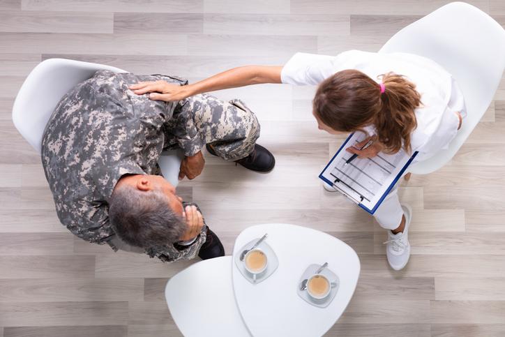 Older man in military uniform sitting in a chair next to a table, holding his head in his hands, being comforted by a female healthcare professional dressed in white who is touching the man's shoulder. Two cups of tea are in front of them on the table.