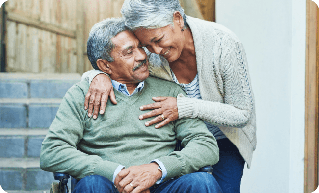 An elderly couple share a side embrace as the husband sits in his wheelchair